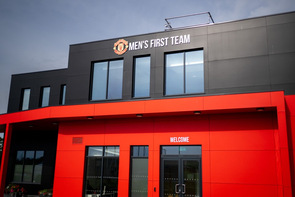 Exterior view of the Manchester United Carrington Training Ground building with "MEN'S FIRST TEAM" and the club logo on a black facade above a red entrance with "WELCOME" over the doors.