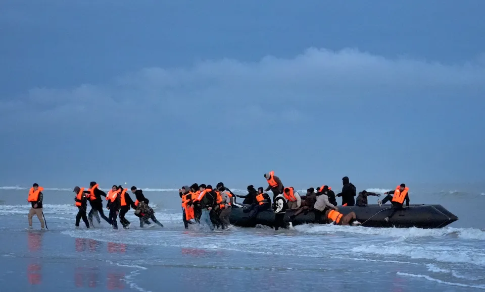 Migrants wearing orange life vests wading into the ocean towards an inflatable boat.