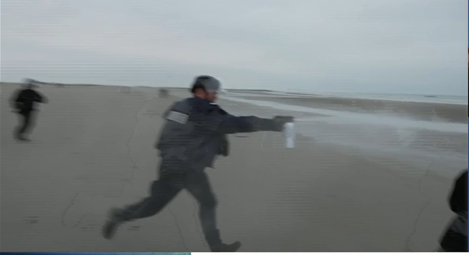 A person in a dark uniform runs along a sandy beach, spraying a white mist from a can toward the right.