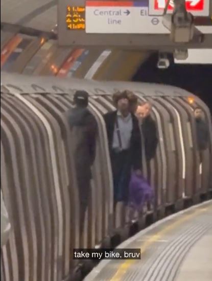 A cyclist standing at a train doorway with their bike next to them, while staff and passengers wait.