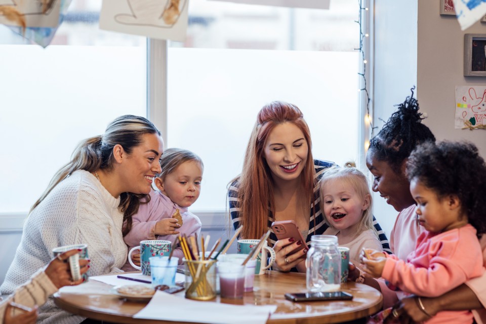 Mothers and toddlers sitting around a table in a preschool, talking, drinking tea, and snacking, with one mother showing something on her smartphone.