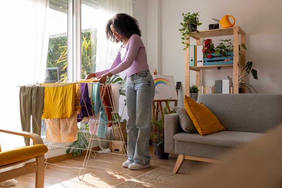 A young woman drying laundry on a clothes rack in a sunlit living room.