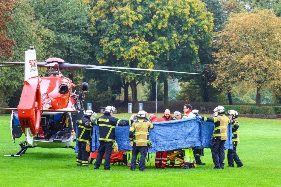 Paramedics and firefighters load a patient on a stretcher into a red helicopter on a grassy field.