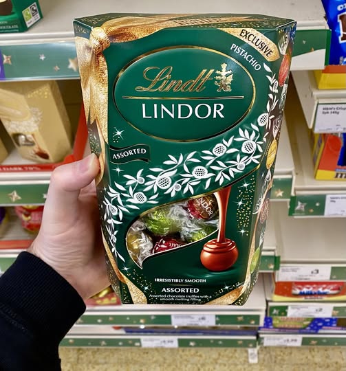 A person holding a box of Lindt Lindor assorted chocolate truffles with an exclusive pistachio flavor.