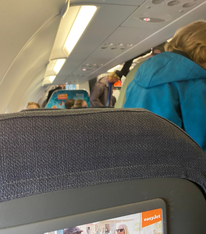 View of the cabin of an EasyJet airplane with passengers standing and an advertisement for EasyJet visible from a seat.