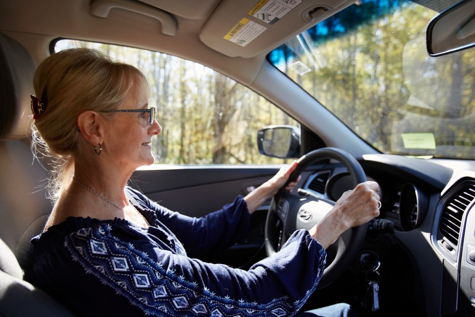 Interior view of a senior woman in glasses smiling while driving a car through a sunlit forest.
