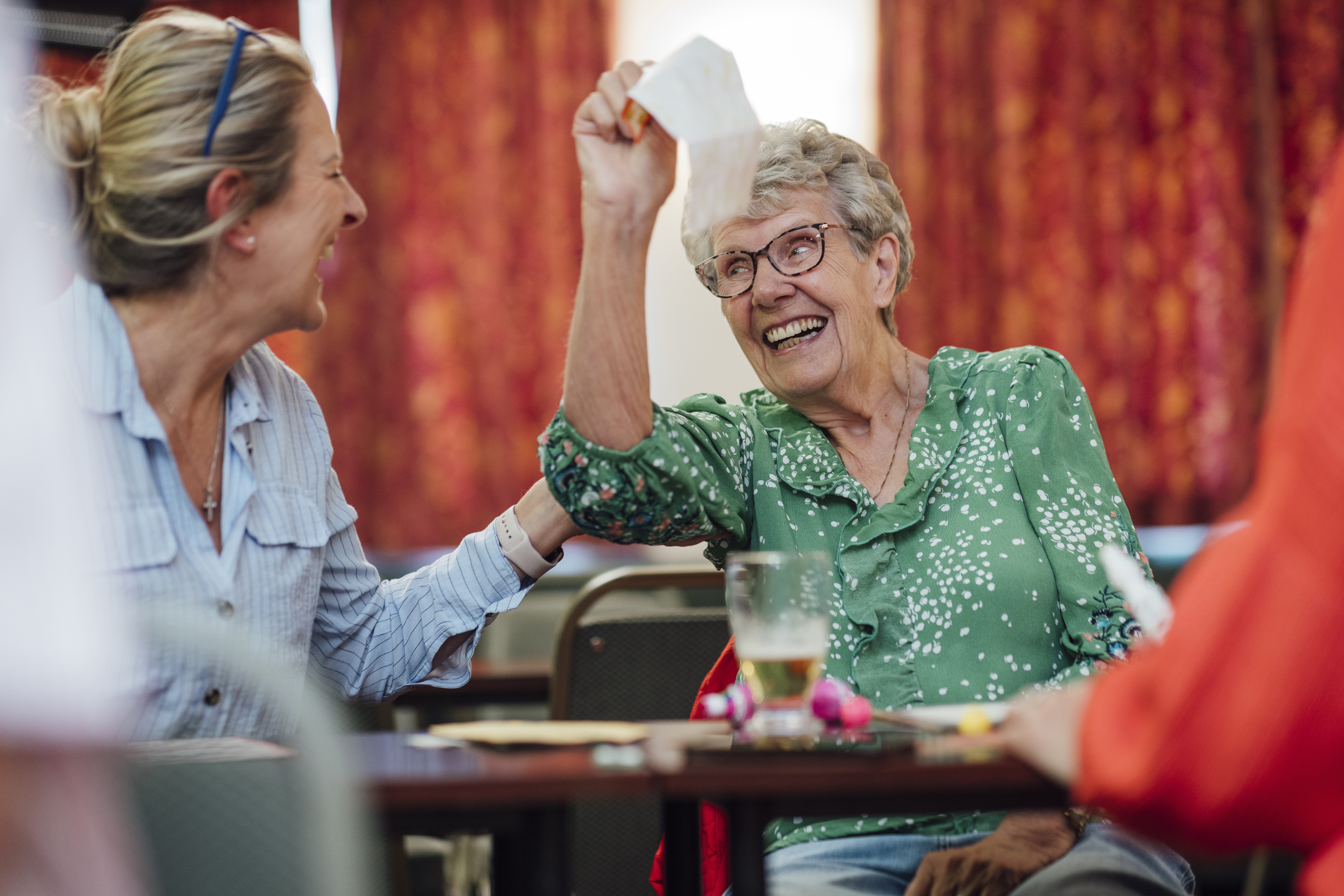 A woman raising a bingo card in the air while laughing, with another woman laughing next to her.