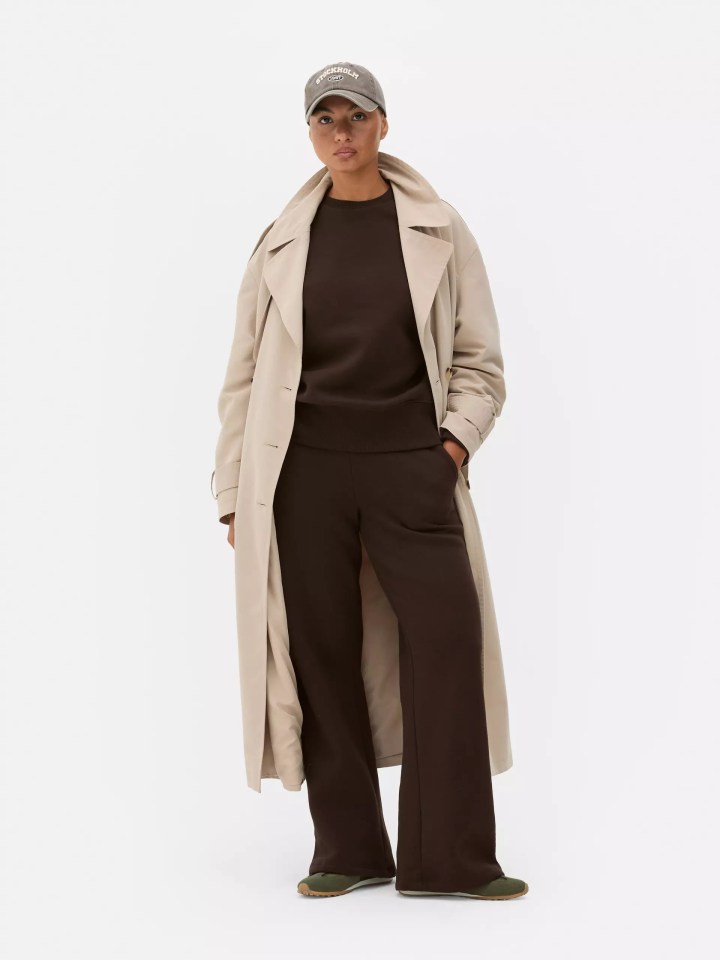 A woman wearing a beige trench coat over a brown tracksuit, green sneakers, and a grey 'Stockholm 1997' cap.