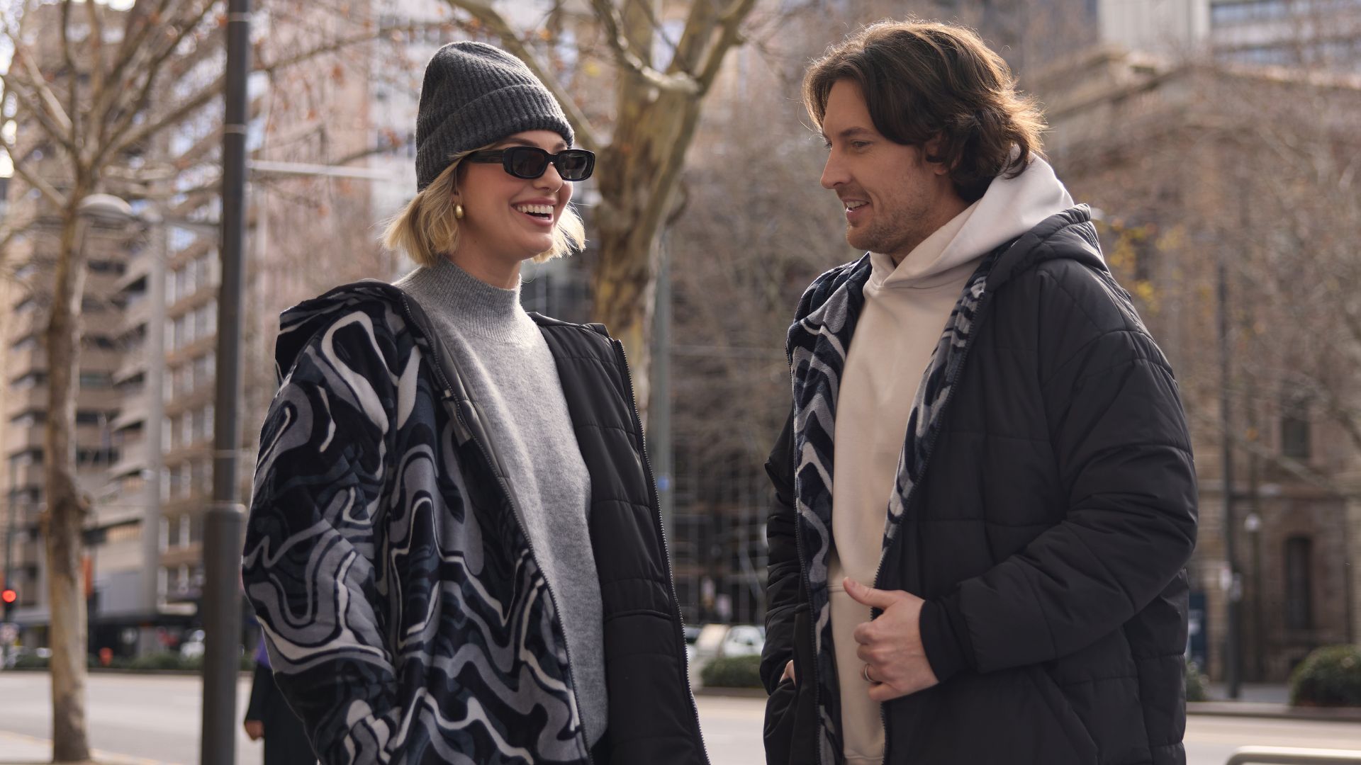 A man and a woman wearing quilted coats, with the woman wearing a beanie and sunglasses.