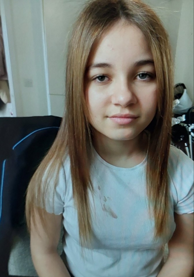 A young woman with long, straight light brown hair and a light blue t-shirt with a stain on the chest, looking directly at the camera.