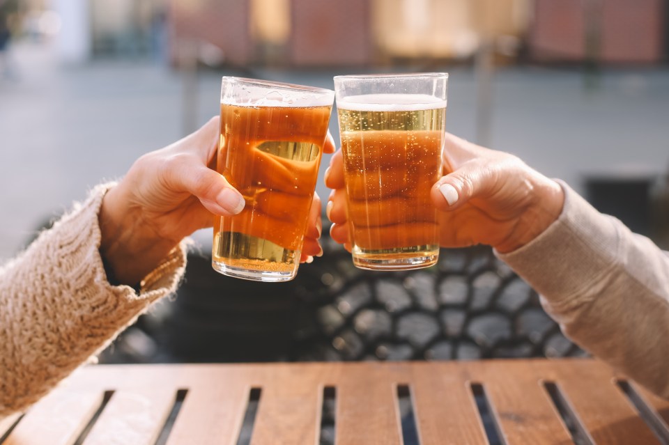 Two people toasting with pint glasses of beer at an outdoor wooden table.