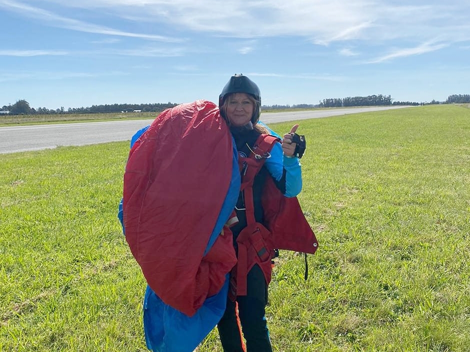 Skydiver Rosana Mabel Back giving a thumbs up, wearing a helmet and blue and red skydiving gear.