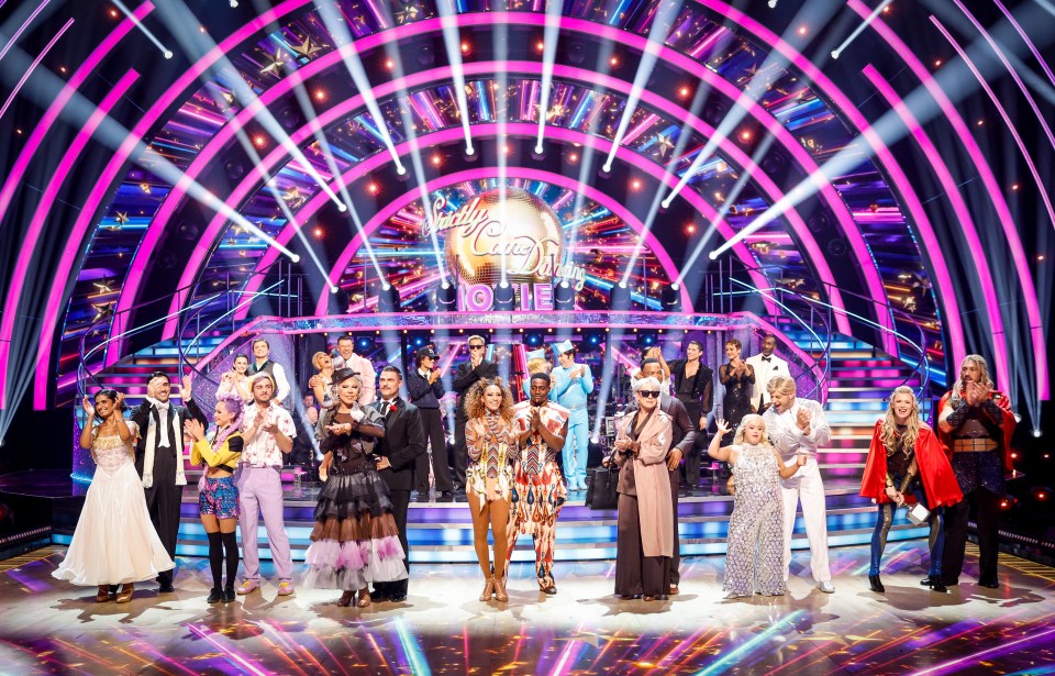 A group of costumed dancers and celebrities standing on a brightly lit dance floor with a large disco ball in the background.