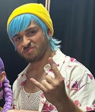 George Clarke in a yellow beanie and blue wig, making a fierce face and rock-and-roll hand gesture.