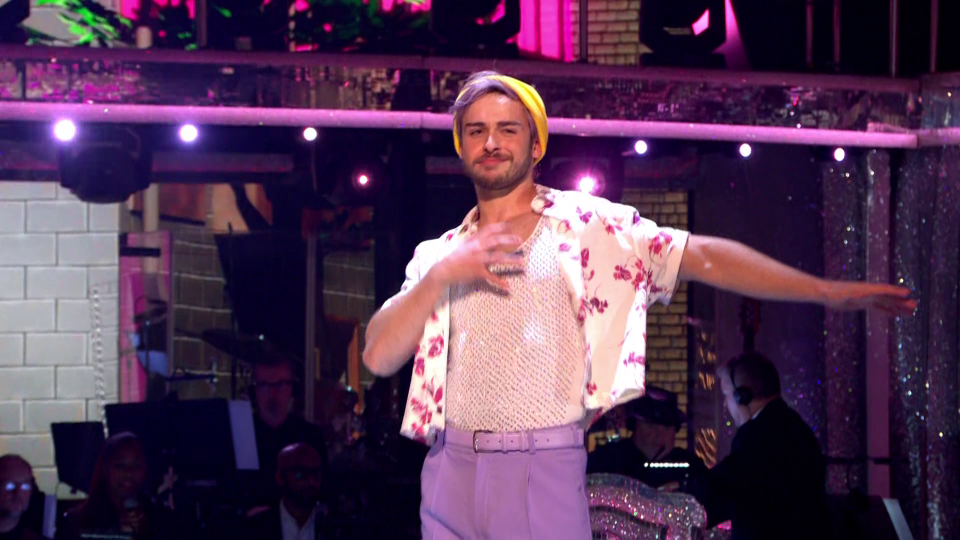 Johannes Radebe in a floral shirt, lilac pants, and a yellow beanie dancing.