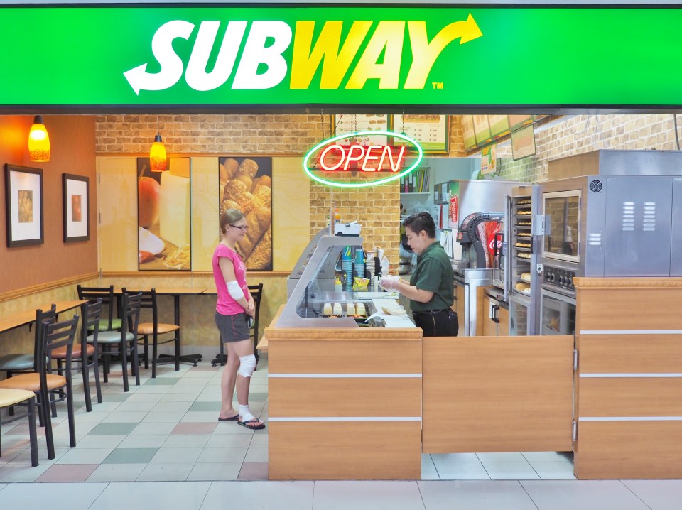 Interior view of a Subway restaurant with a customer at the counter and an employee preparing a sandwich.