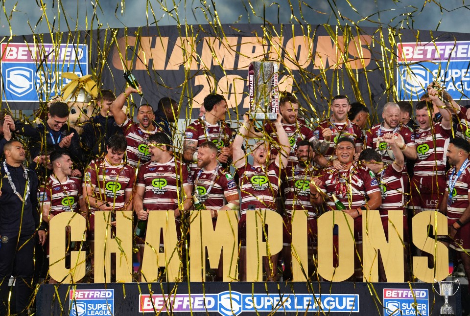 Wigan Warriors players celebrating with the trophy, with gold confetti falling and a "CHAMPIONS" sign in front.