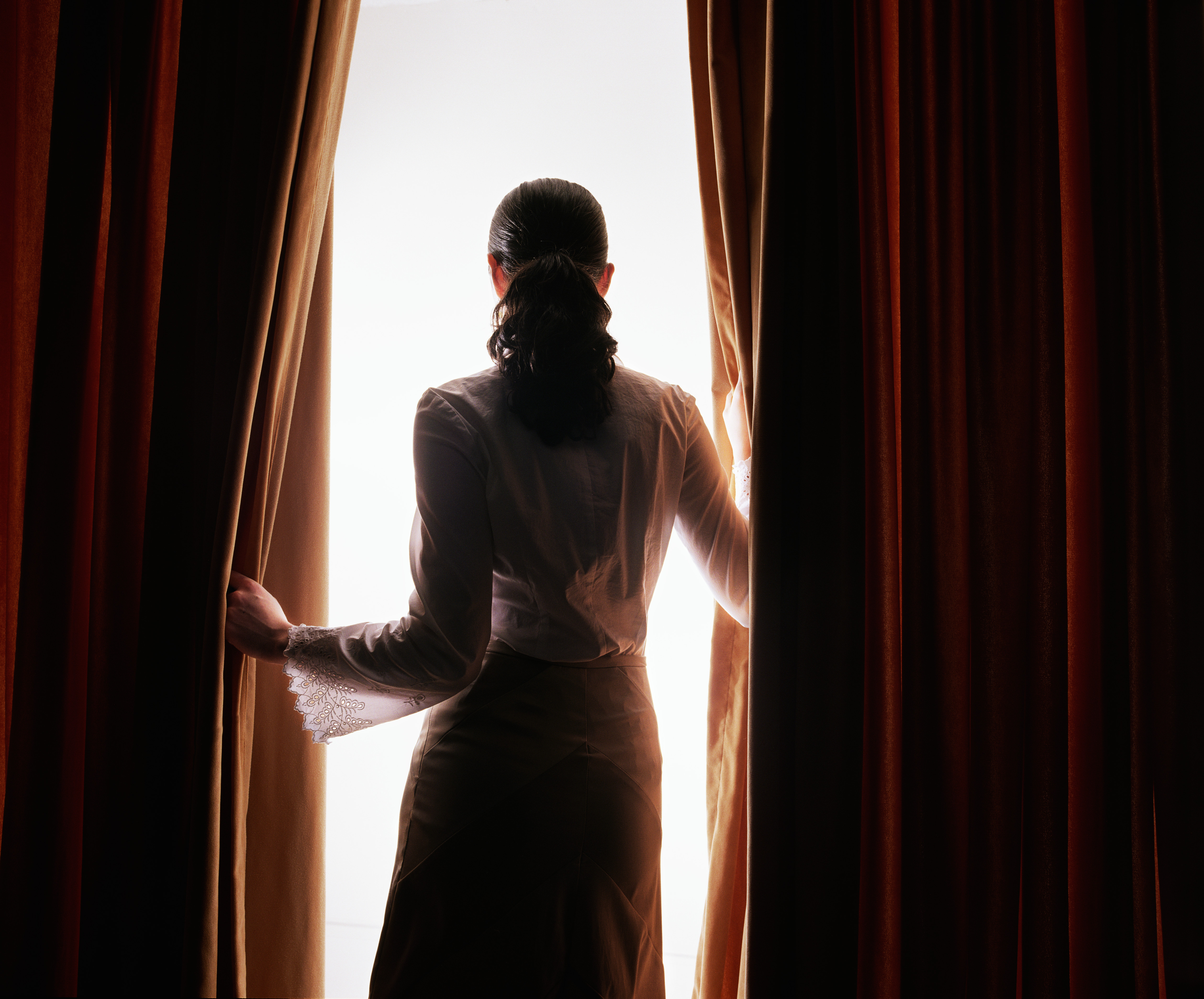 Woman with a ponytail pulling back red curtains to reveal a bright light.