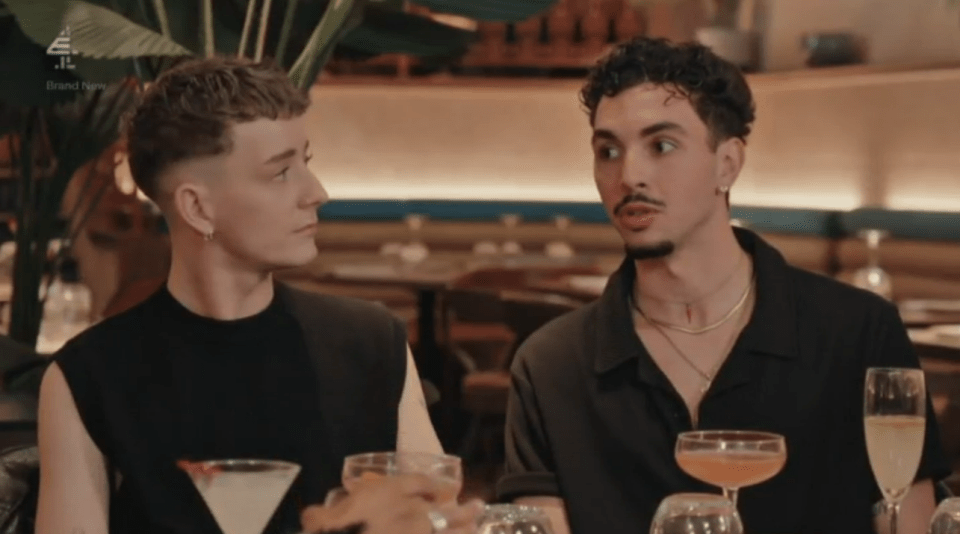 Two men at a table with drinks, one speaking, the other listening.