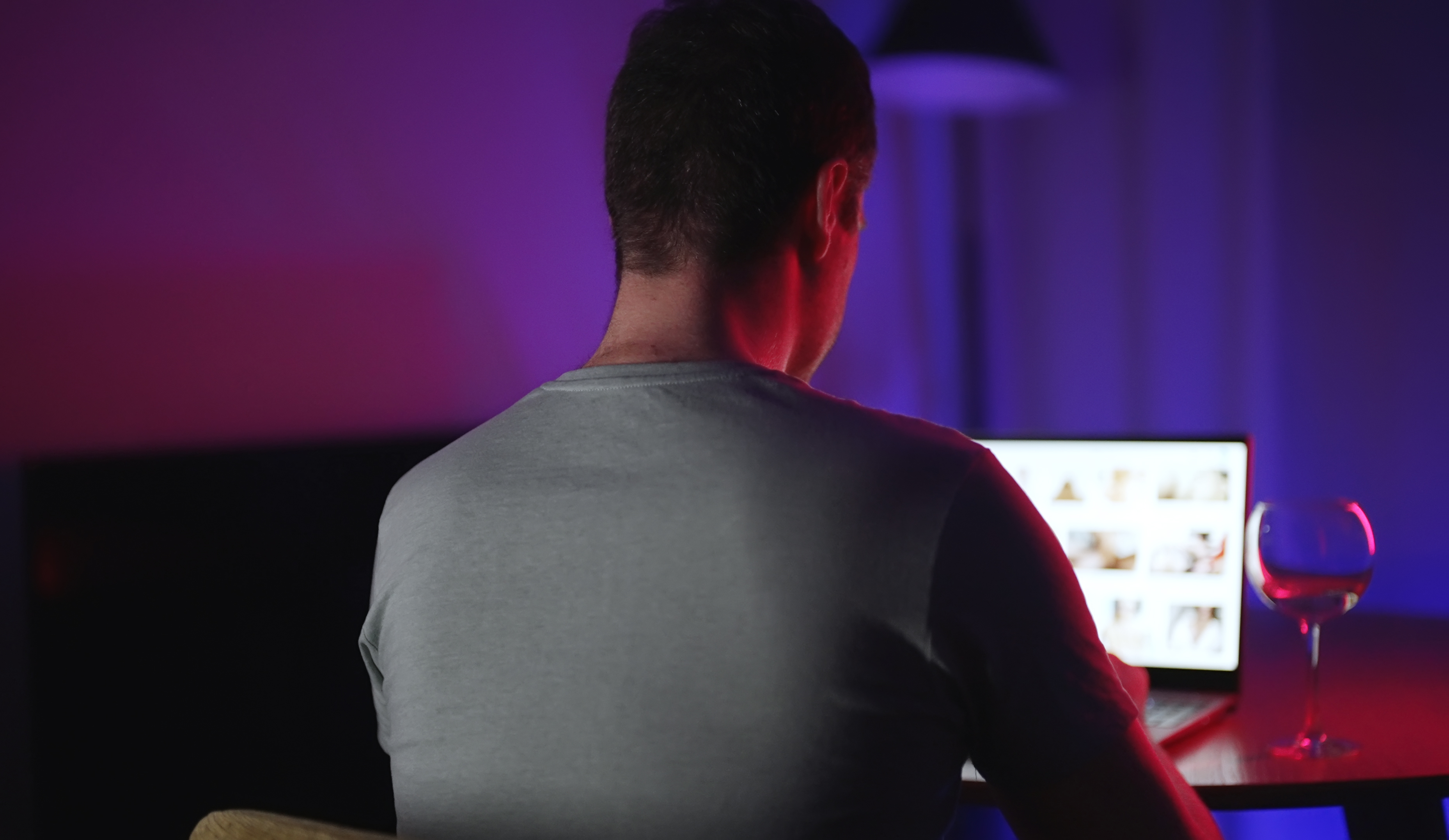 A man watches adult content on a laptop at night, a glass of wine beside him.
