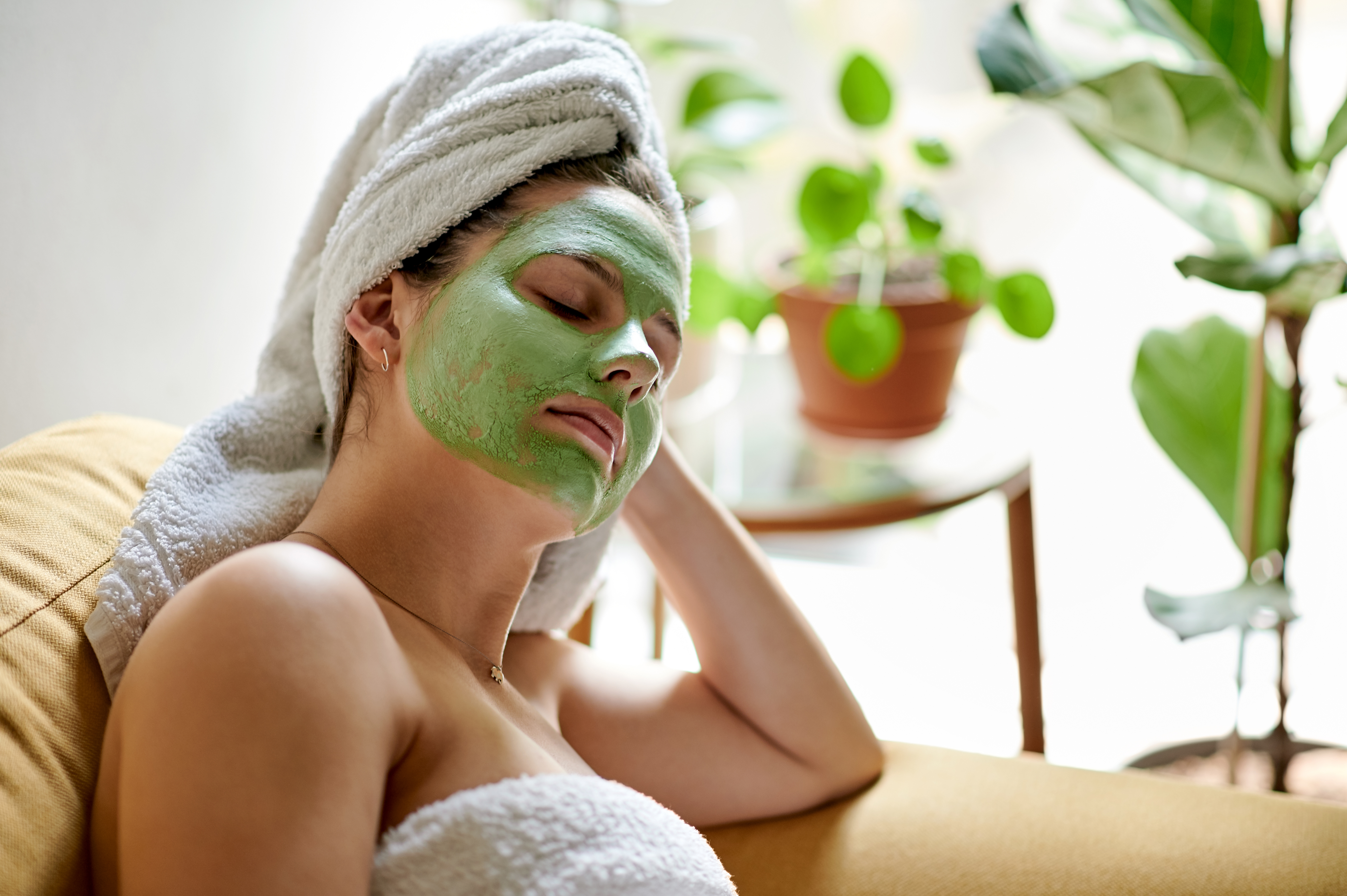 Woman with a green face mask and a white towel on her head, relaxing with her eyes closed.