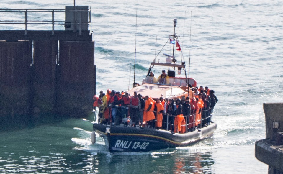 An RNLI lifeboat carrying many migrants in orange and yellow life vests approaches a dock.
