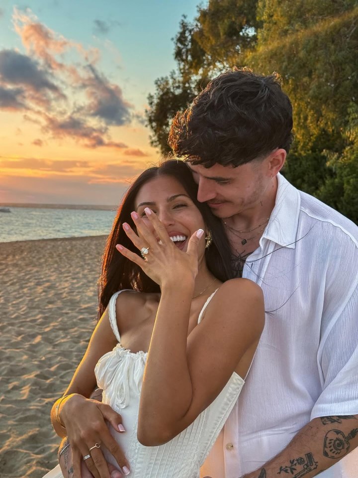 A woman showing off an engagement ring while a man embraces her from behind at sunset on a beach.