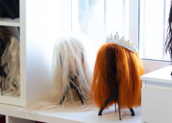 Wigs of various colors and styles, including a tiara, sit on a white shelf in a bright dressing room, likely belonging to a performer or entertainer.