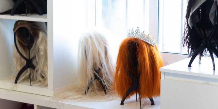 Wigs of various colors and styles, including a tiara, sit on a white shelf in a bright dressing room, likely belonging to a performer or entertainer.