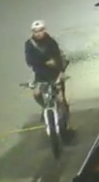 Security camera footage of a person in a baseball cap, dark jacket, and light pants riding a motorcycle.