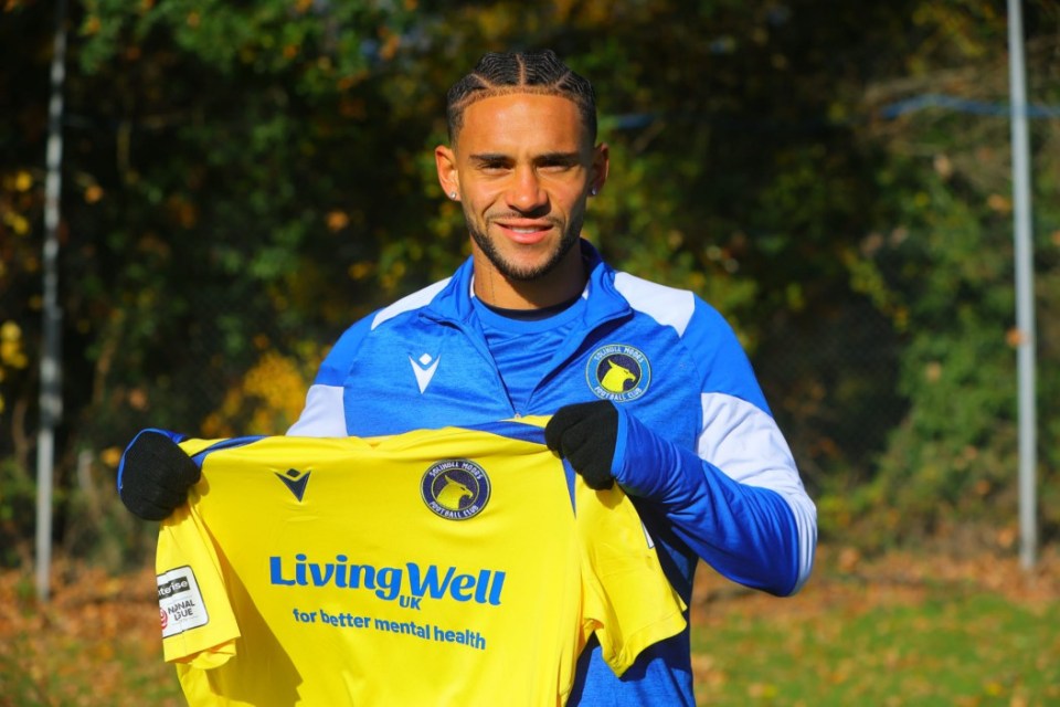 A man in a blue and white long-sleeved shirt holding a yellow soccer jersey with "LivingWell UK for better mental health" on it.