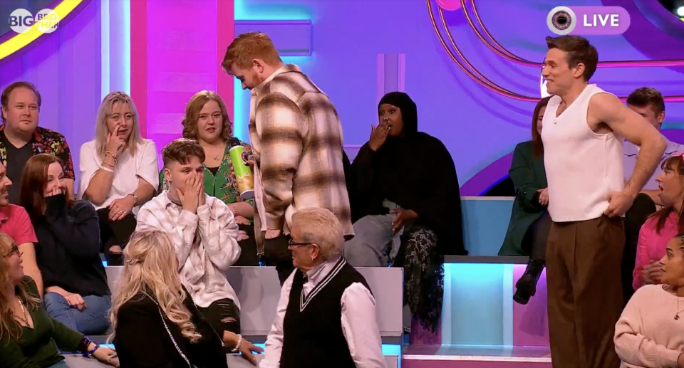 An audience watches a man on stage in a tank top and brown pants, with two others behind him on a purple and pink stage.