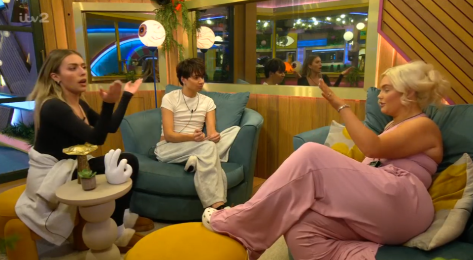 Three people talking in a living room, two women gesturing while a third person listens.