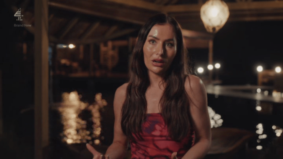 A woman in a red and black dress speaking indoors at night, with lights reflecting on water behind her.