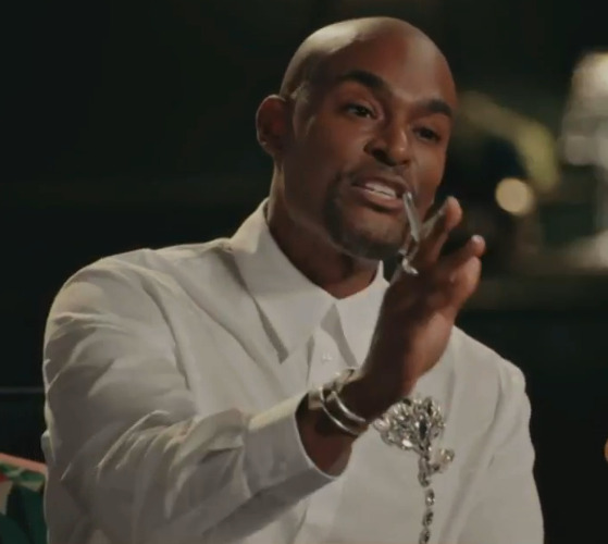 A man wearing a white shirt and a brooch with a chain, gesturing with his hand.