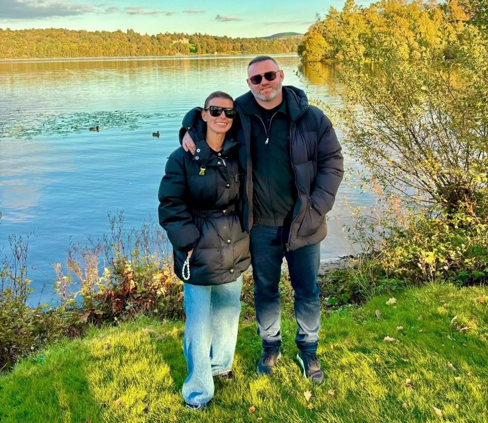 Wayne and Coleen Rooney standing together outdoors next to a lake with trees in the background.