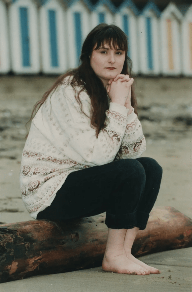 Woman crouching barefoot on a log, with beach huts in the background.