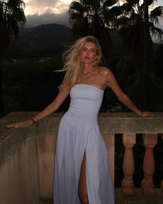 Alica Schmidt leaning on a stone balcony railing, wearing a light blue dress with a high slit, with palm trees and mountains in the background.