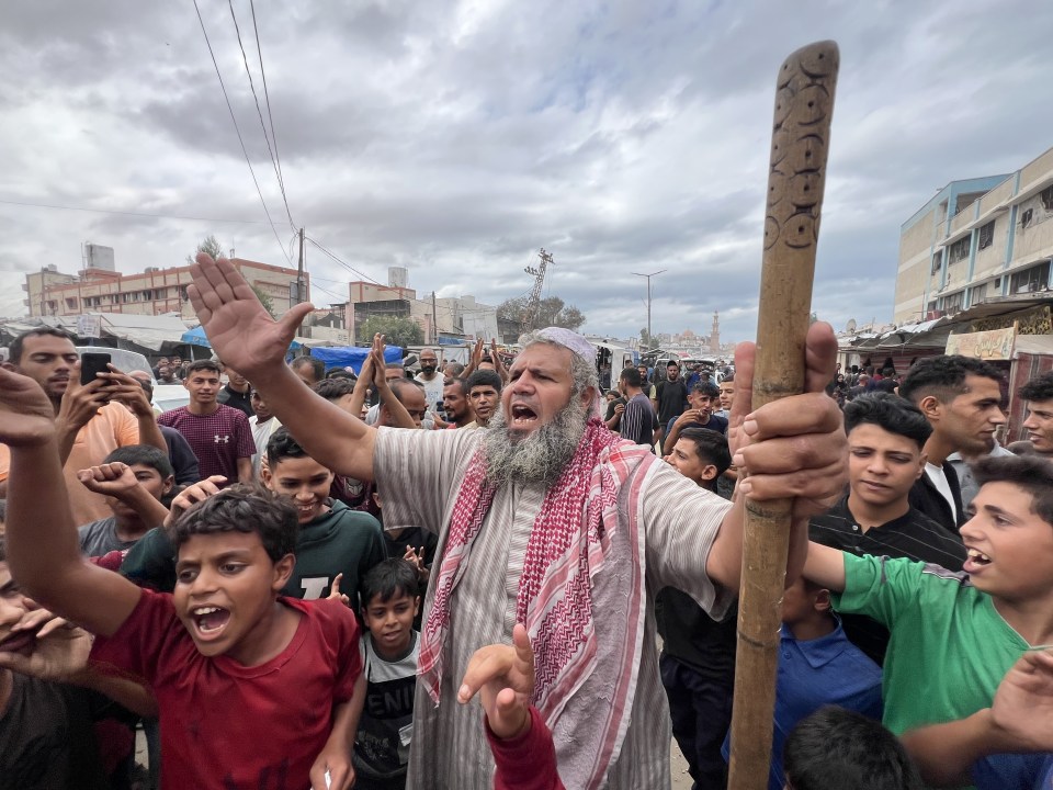 A man with a white beard and head covering shouting, surrounded by a crowd of men and boys, some raising their hands.