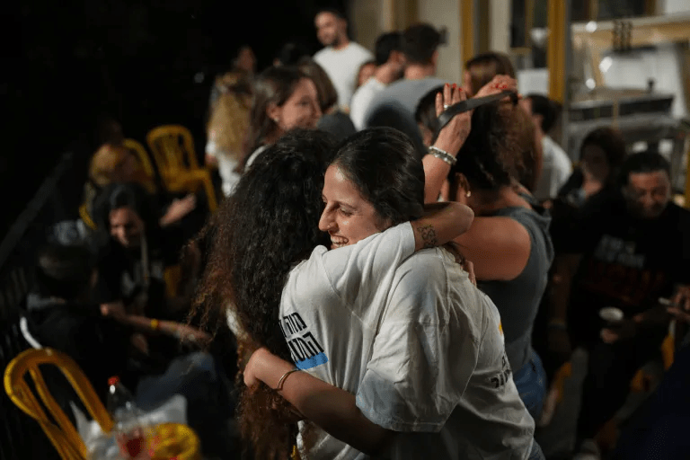 Two women hugging in a crowd, one in a white shirt and the other with dark, curly hair.