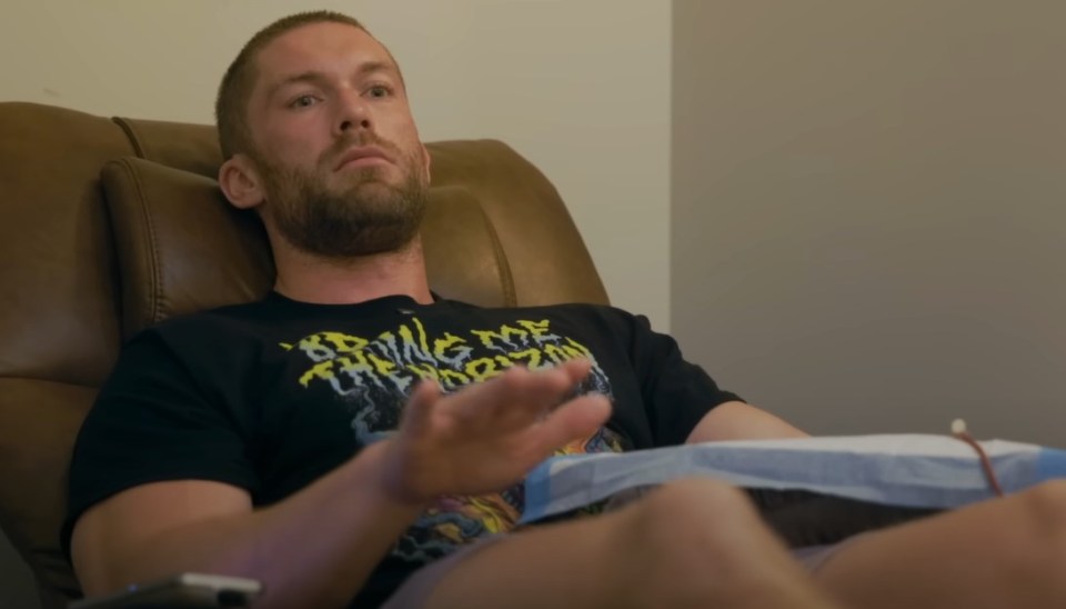 Chris Williamson reclines in a brown leather chair, wearing a black t-shirt, with medical equipment attached to his arm.