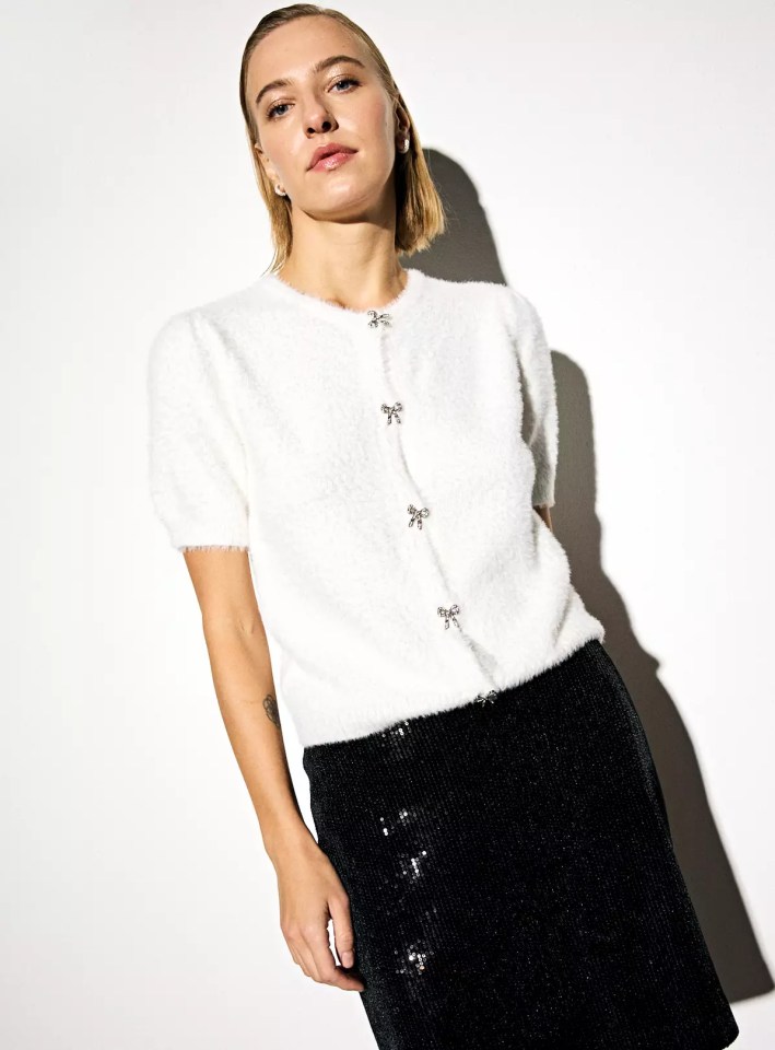 A woman wearing a white fuzzy short-sleeved cardigan with bow-shaped clasps and a black sequined skirt.