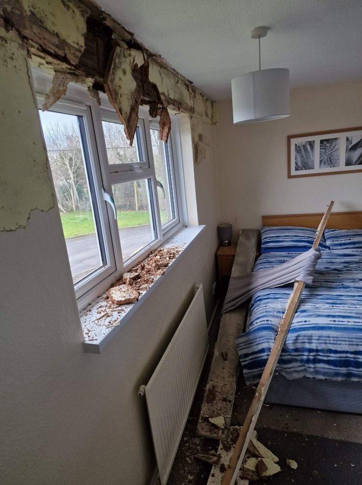 A damaged bedroom with a broken window frame, debris on the windowsill and floor, and wood planks on the bed.