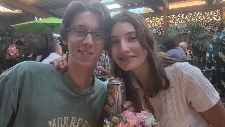 A couple poses at an outdoor restaurant, the woman holding a can and a bouquet of flowers.