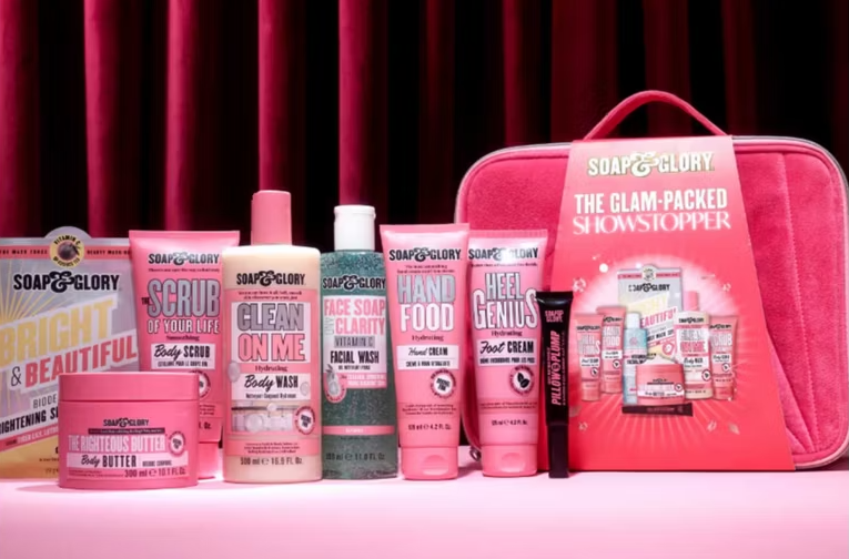 An array of pink Soap & Glory beauty products including body wash, scrub, hand cream, foot cream, and body butter with a pink beauty bag.
