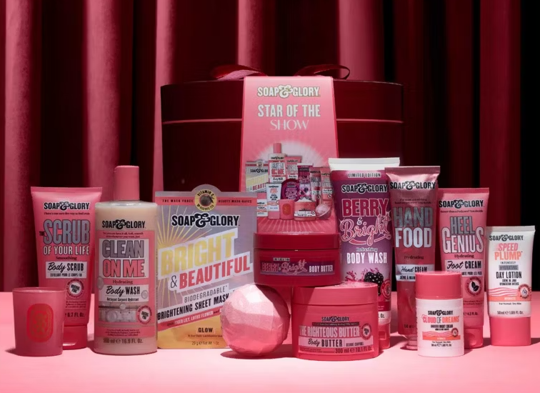 A collection of Soap & Glory products, including body wash, body scrub, body butter, hand cream, foot cream, sheet mask, and day lotion, with a gift box that says "Star of the Show."