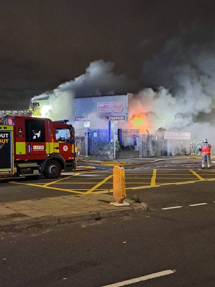 A fire engine and firefighters combating a large fire at an industrial unit at night, with significant smoke and flames visible.