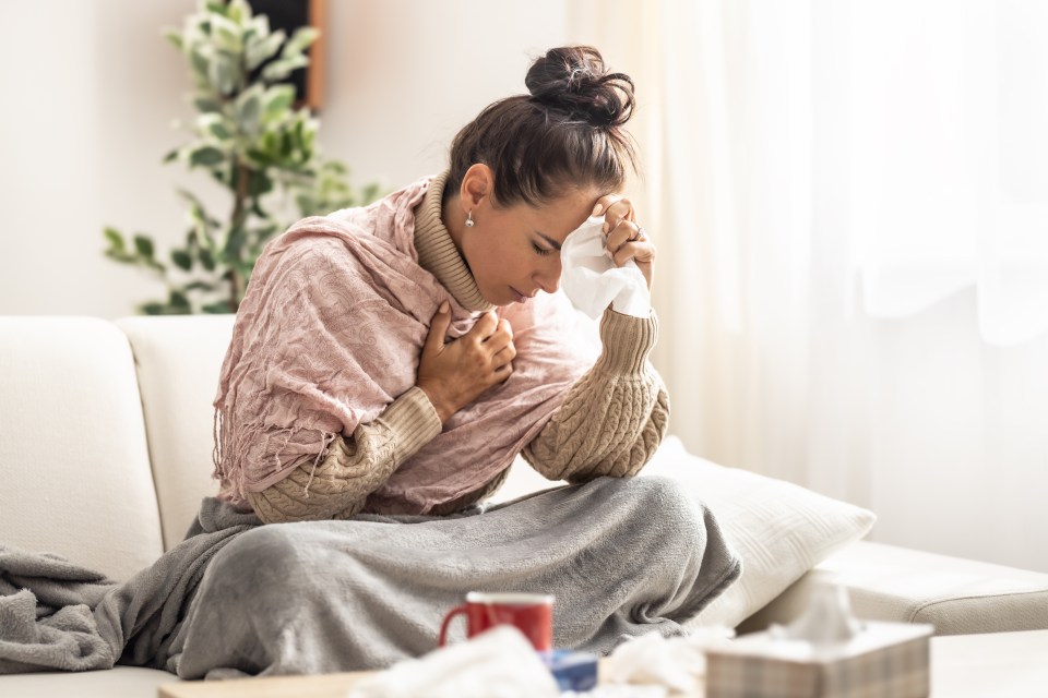 A very sick young woman sitting on a couch, holding her forearm to her forehead and a handkerchief to her nose.