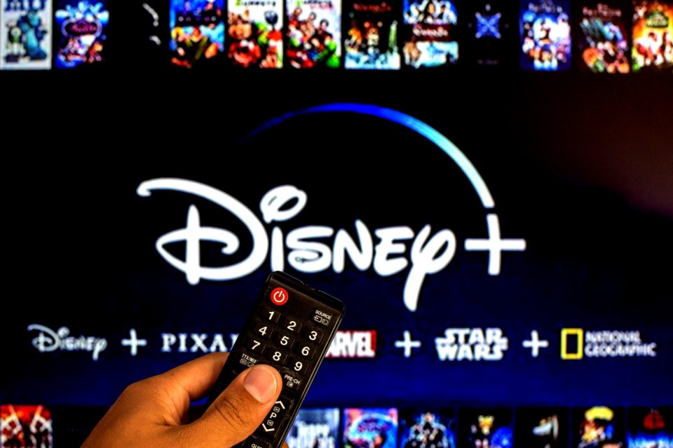 A hand holding a TV remote control in front of a screen displaying the Disney+ logo with other movie titles in the background.