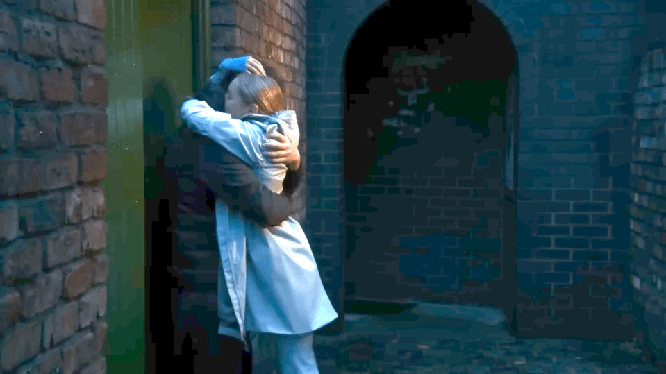 A person in a light-colored coat is hugging another person in a dark coat, standing between a brick wall and a dark archway.